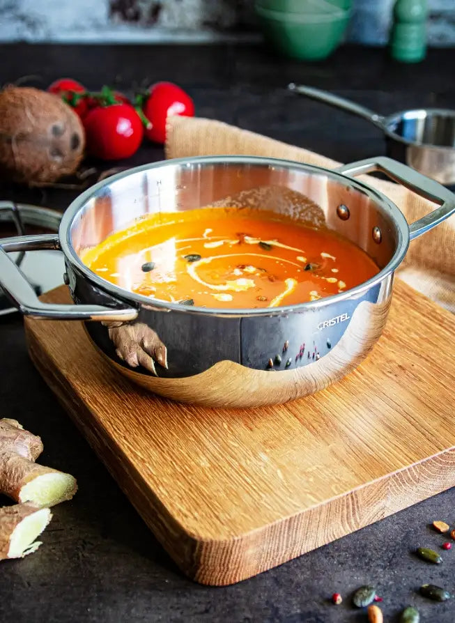 A stainless steel pot filled with orange soup, placed on a wooden cutting board with kitchen ingredients like tomatoes, ginger, and spices around it.