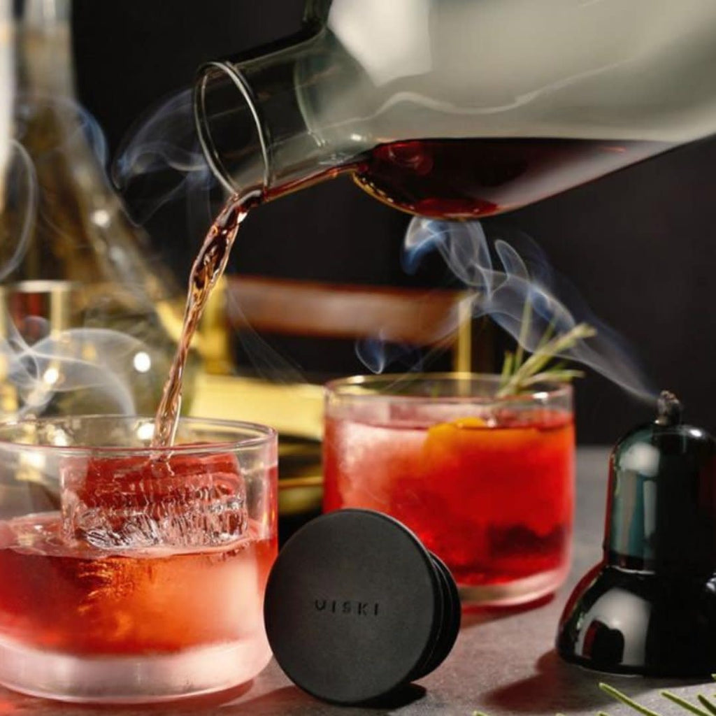 Cocktail being poured with smoke effect into a glass on a bar counter.